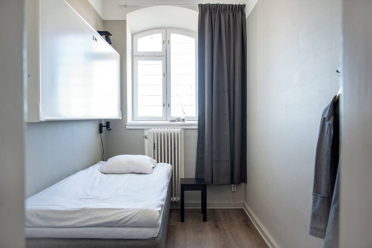 A small, single-bed room with a window, radiator, and a hanging garment.