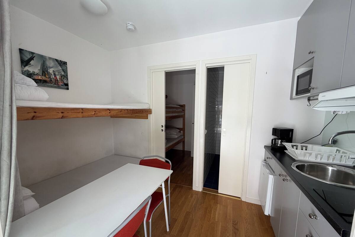 A compact room with a bunk bed, a small dining table with a chair, a kitchen area with a sink and microwave, and a door leading to a bathroom.
