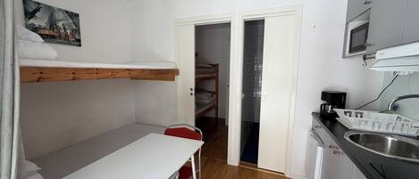 A compact room with a bunk bed, a small dining table with a chair, a kitchen area with a sink and microwave, and a door leading to a bathroom.