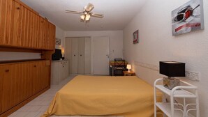 Ein Schlafzimmer mit einem Bett, einem Holzschrank, einem Deckenventilator, weißen Wänden und einem kleinen Tisch mit einer Lampe.