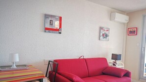 Ein kompakter Wohnraum mit einem roten Sofa, einem kleinen Esstisch mit gestreiftem Tischläufer und einem Stuhl. Zwei Wandklimageräte und gerahmte Kunstwerke zieren die Wände.