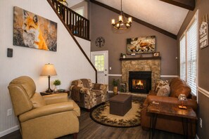 A living room with a fireplace, leather sofas, a patterned armchair, a staircase, and a chandelier.