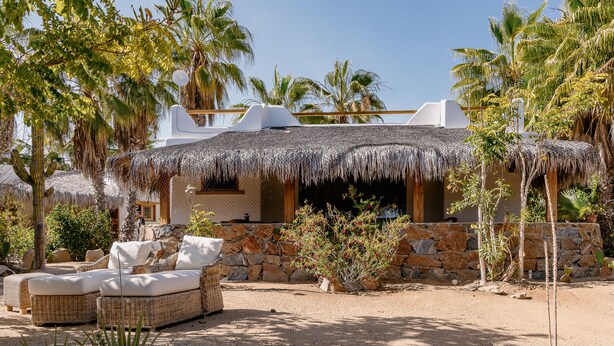 A thatched-roof building with stone walls, surrounded by palm trees and a sandy area with a wicker sofa and cushions.