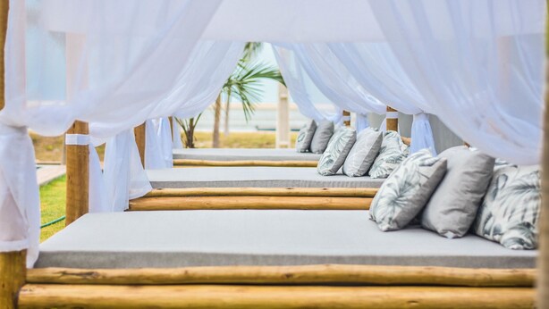 A row of wooden benches with white cushions, draped with white fabric, leading to a building. - Locanda Dei Venti (Itarema)