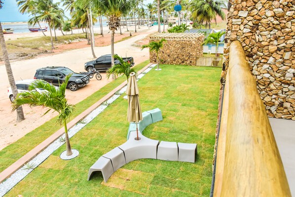 Un césped cuidado con un espacio de descanso singular, un muro de piedra y vistas a la playa con coches aparcados y barcos.