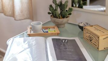 Una cama con colchón blanco, un libro, una taza, una bandeja con una taza y un paquete, un cactus en maceta y un caja de madera sobre una mesa de vidrio.