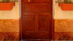 A wooden door with a metal decorative arch above it, flanked by two orange flower pots. A sign with the number "100" is visible on the wall to the left.