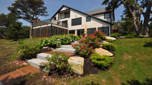 Ett modernt hus i två våningar med en balkong, omgivet av en välskött trädgård med stenplattor och blommande växter.