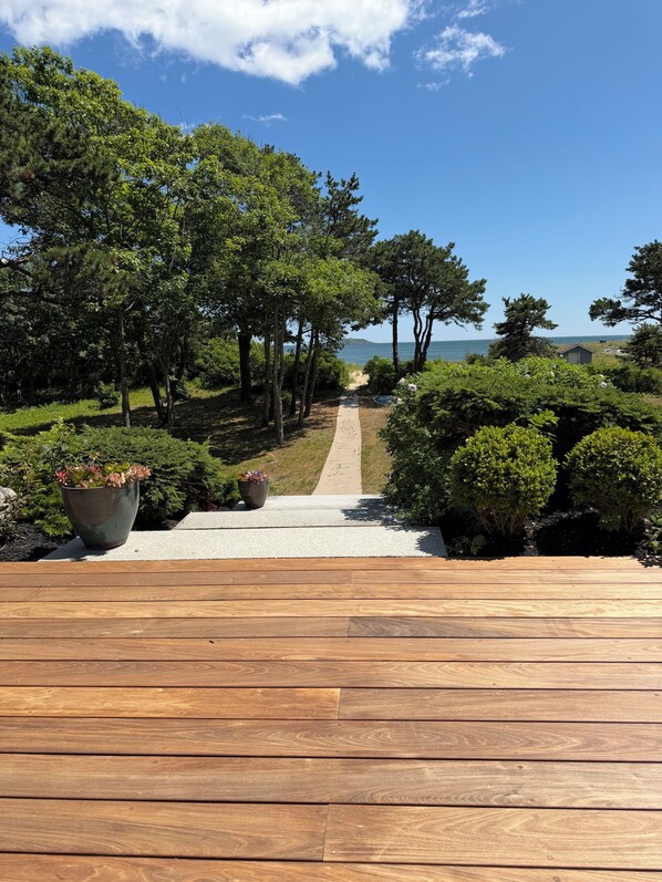 A wooden deck leading to a pathway surrounded by trees and shrubs, with a view of the sea in the distance.