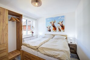 A bedroom with a wooden bed, a window with a view of a brick building, a painting of cows, and a wooden wardrobe.