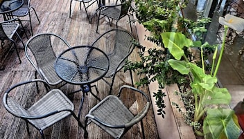 Una terraza de madera con sillas y mesas de metal, rodeada de follaje.