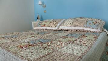 A bedroom with a floral quilted bedspread, a nightstand with a lamp, and a wall-mounted clock.