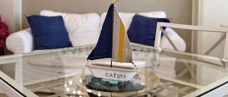 Un velero modelo sobre una mesa de vidrio con una base decorativa, un sofá blanco con cojines azules y un espejo con marco floral al fondo.