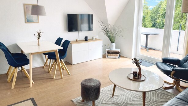 A modern living room with a dining area, a flat-screen TV mounted on the wall, a balcony with a table and chairs, and a large window offering a view of greenery.