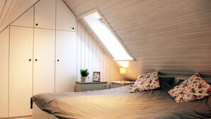 A bedroom with a sloped ceiling, a bed with floral pillows, a nightstand with a lamp, and a small plant.