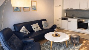 A modern living room with a black leather sofa, a round coffee table, and a kitchen area with white cabinets and a built-in oven.