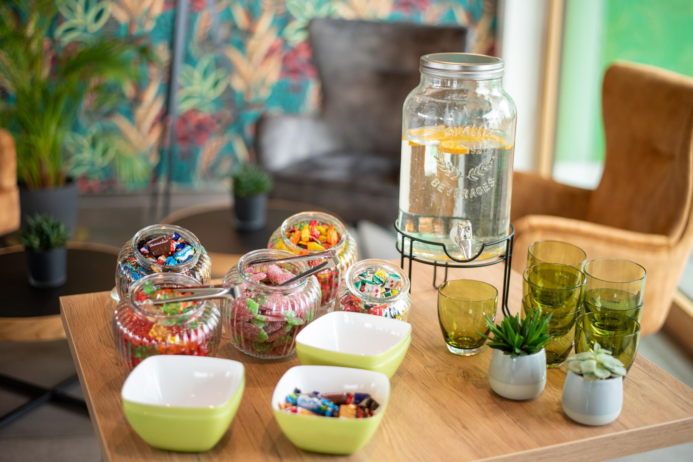 A wooden table with jars of colorful candies, a glass jar with a yellow drink, and small potted plants.