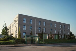 A modern hotel building with a flat roof, multiple windows, and a covered outdoor seating area.