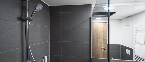 Un baño moderno con paredes de azulejos oscuros, mampara de ducha de vidrio y una puerta de madera visible en el reflejo.