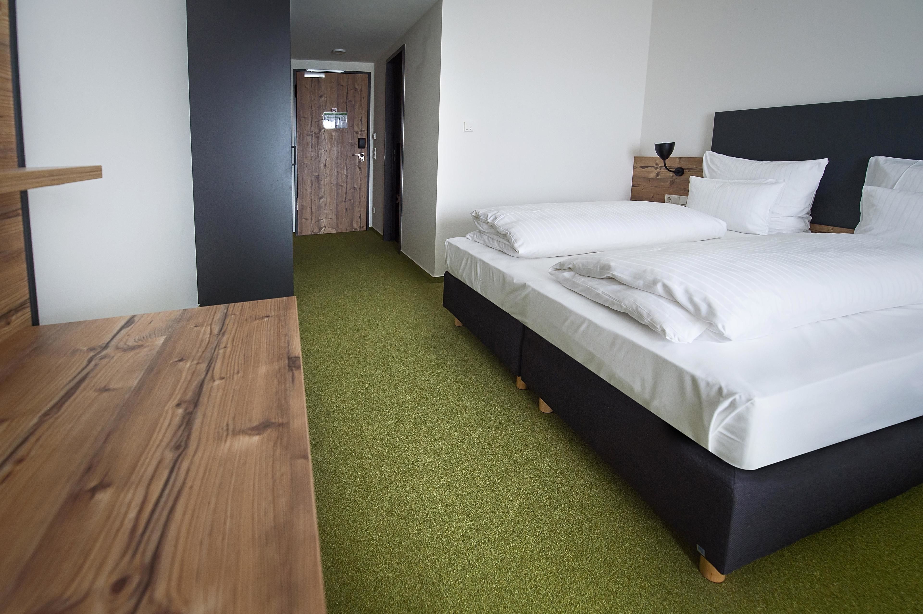 Hotellihuone, jossa on puinen työpöytä, valkoisilla lakanoilla varustettu sänky ja vihreä matto.