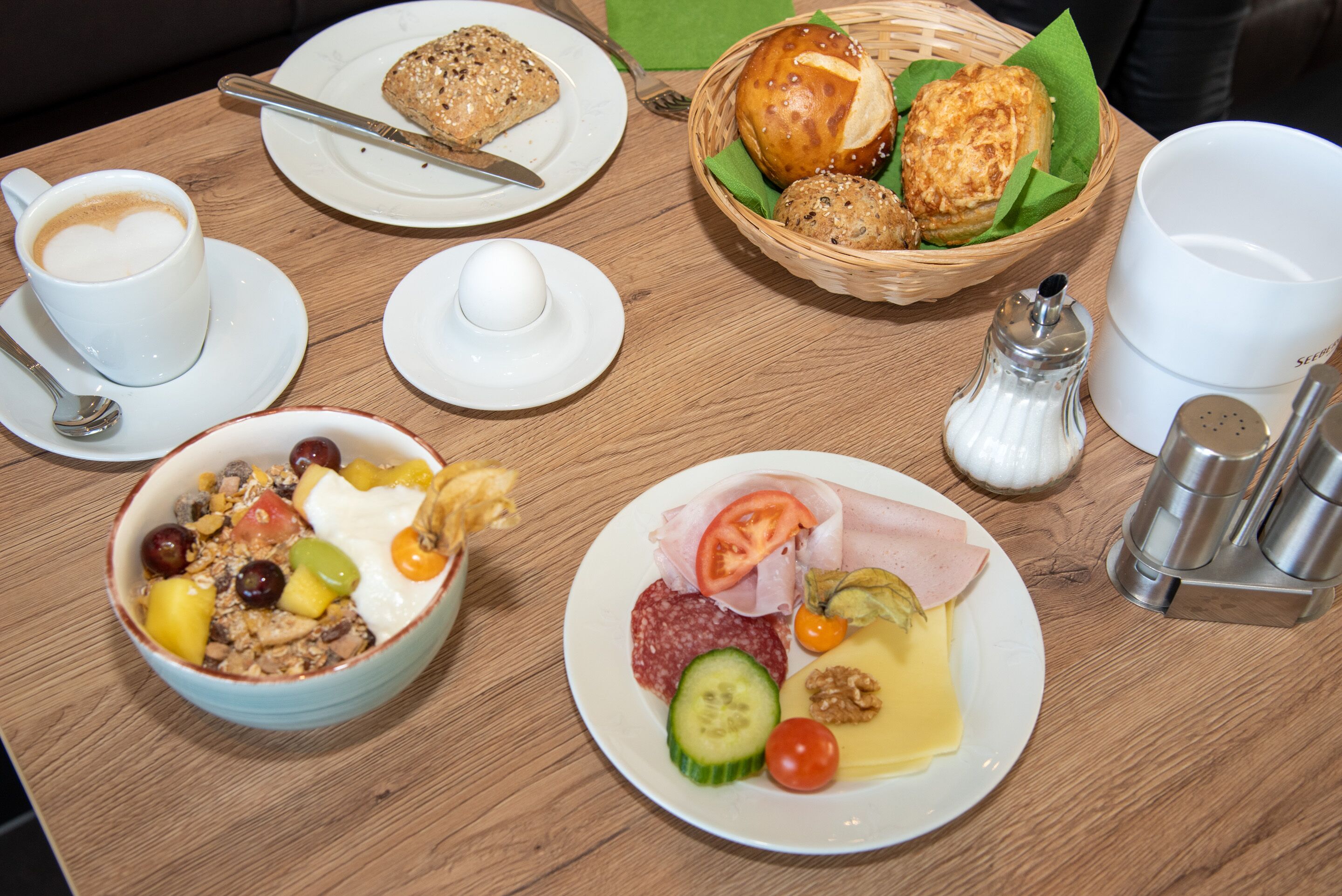 A breakfast table with a cup of coffee, a bowl of cereal, a plate with ham, cheese, and vegetables, and a basket with bread and pastries.