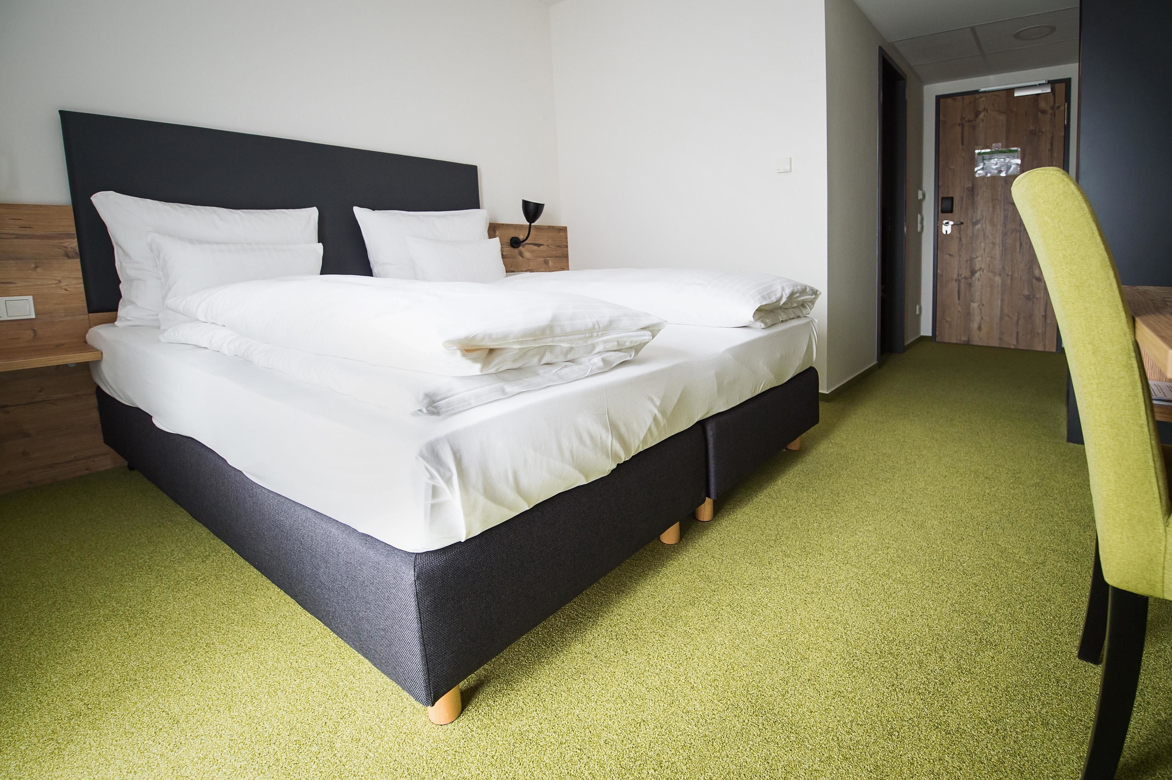 Una cama bien hecha con sábanas blancas, un cabecero de madera y una mesita de noche con una lámpara. La habitación tiene una alfombra verde y una puerta de madera al fondo.