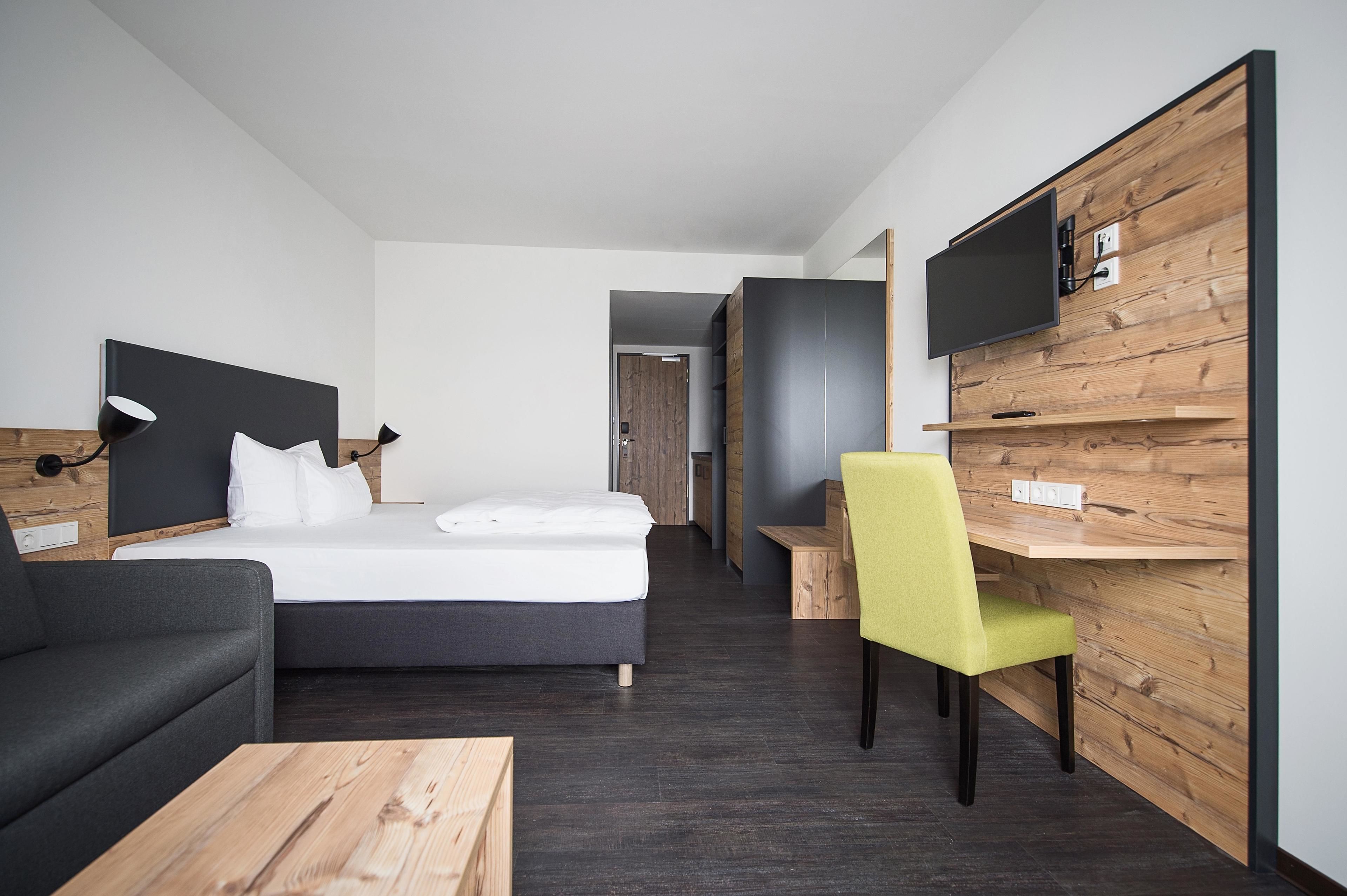 Ein modernes Hotelzimmer mit Bett, Schreibtisch, Stuhl, Fernseher an einer Holzwand und einer Couch.