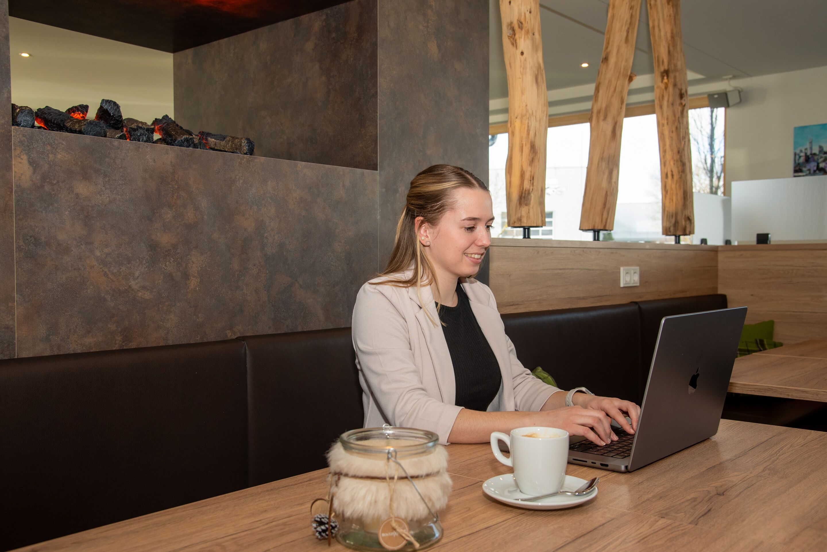 Una persona sentada a una mesa de madera con una computadora portátil, una taza de café y una vela en un frasco.