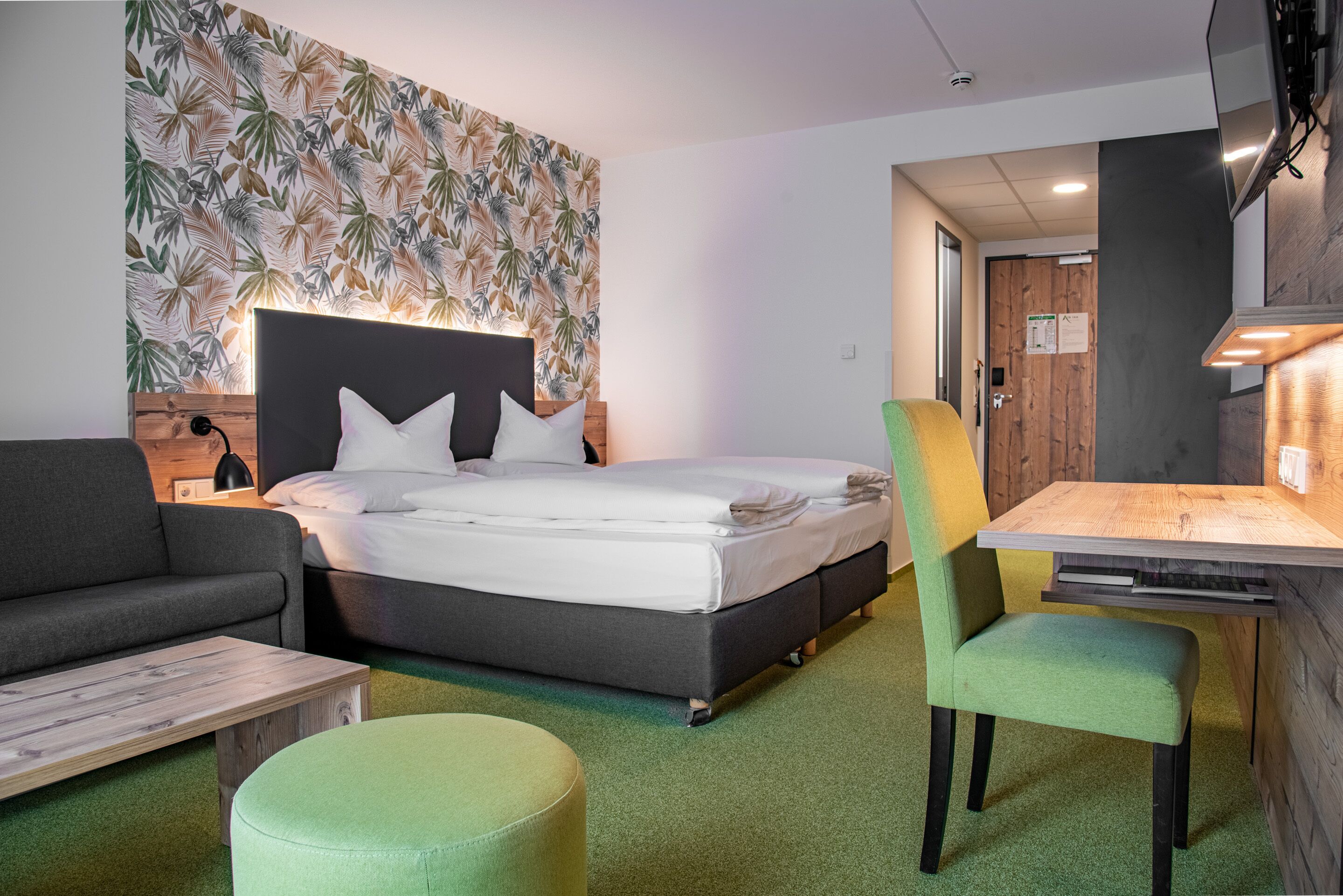 A modern hotel room with a bed, a sofa, a dining table, and a green ottoman.