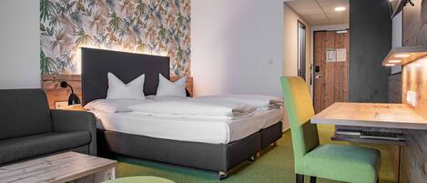 A modern hotel room with a bed, a sofa, a dining table, and a green ottoman.