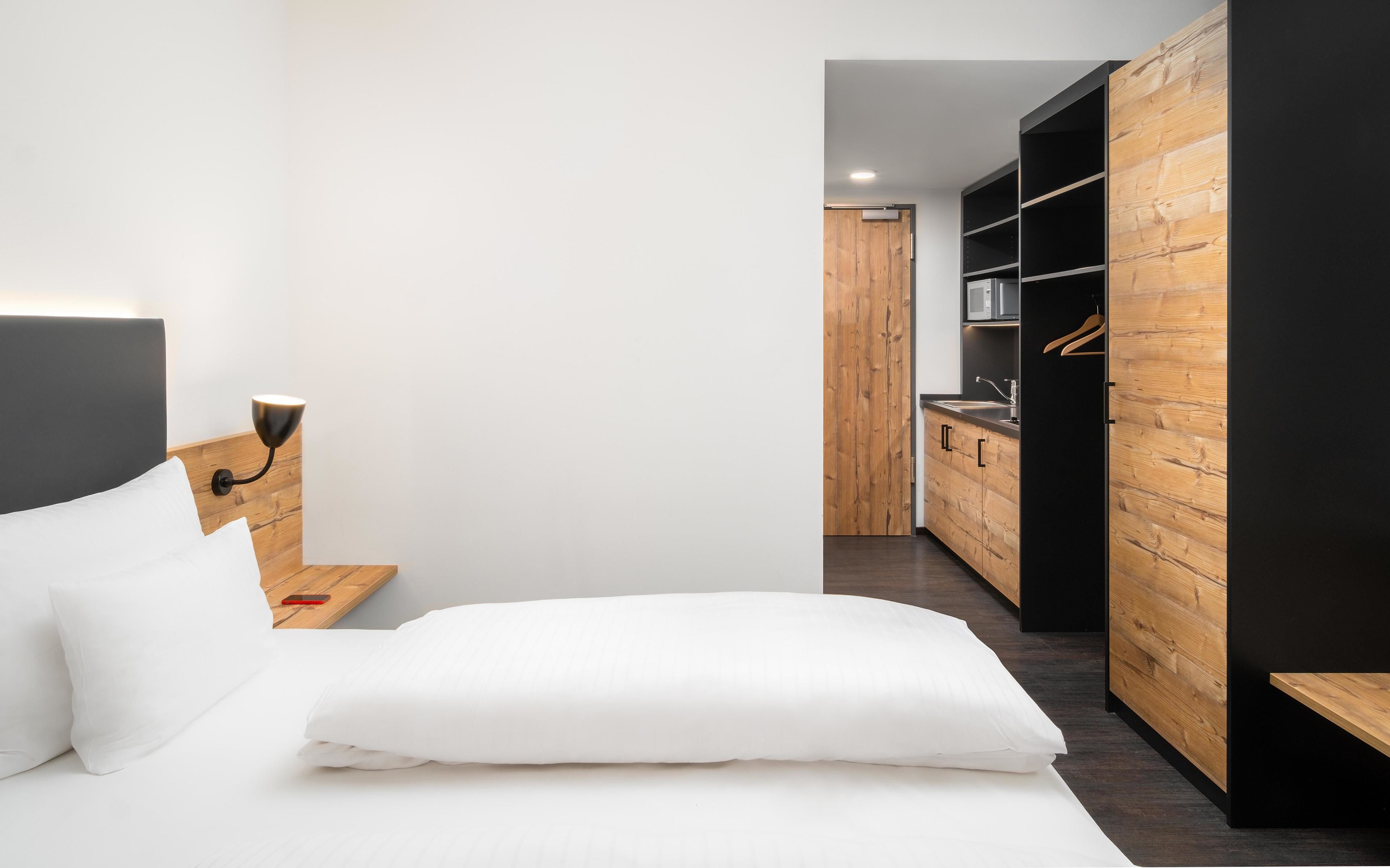 Una habitación de hotel moderna con una cama, una mesita de noche, un armario de madera y una kitchenette.