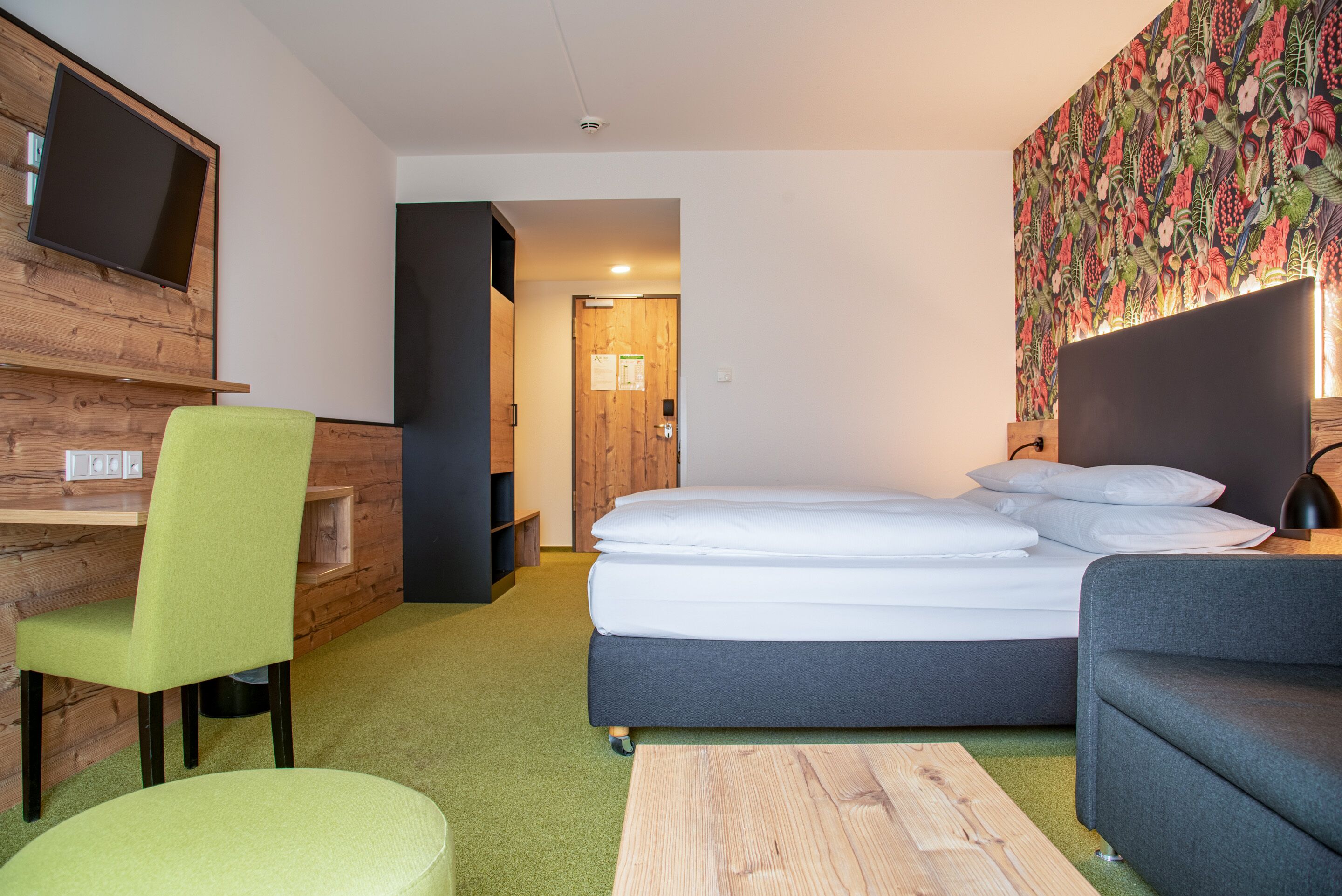 Un hotel moderno con cama, escritorio, silla verde, sofá gris y televisor montado en la pared.