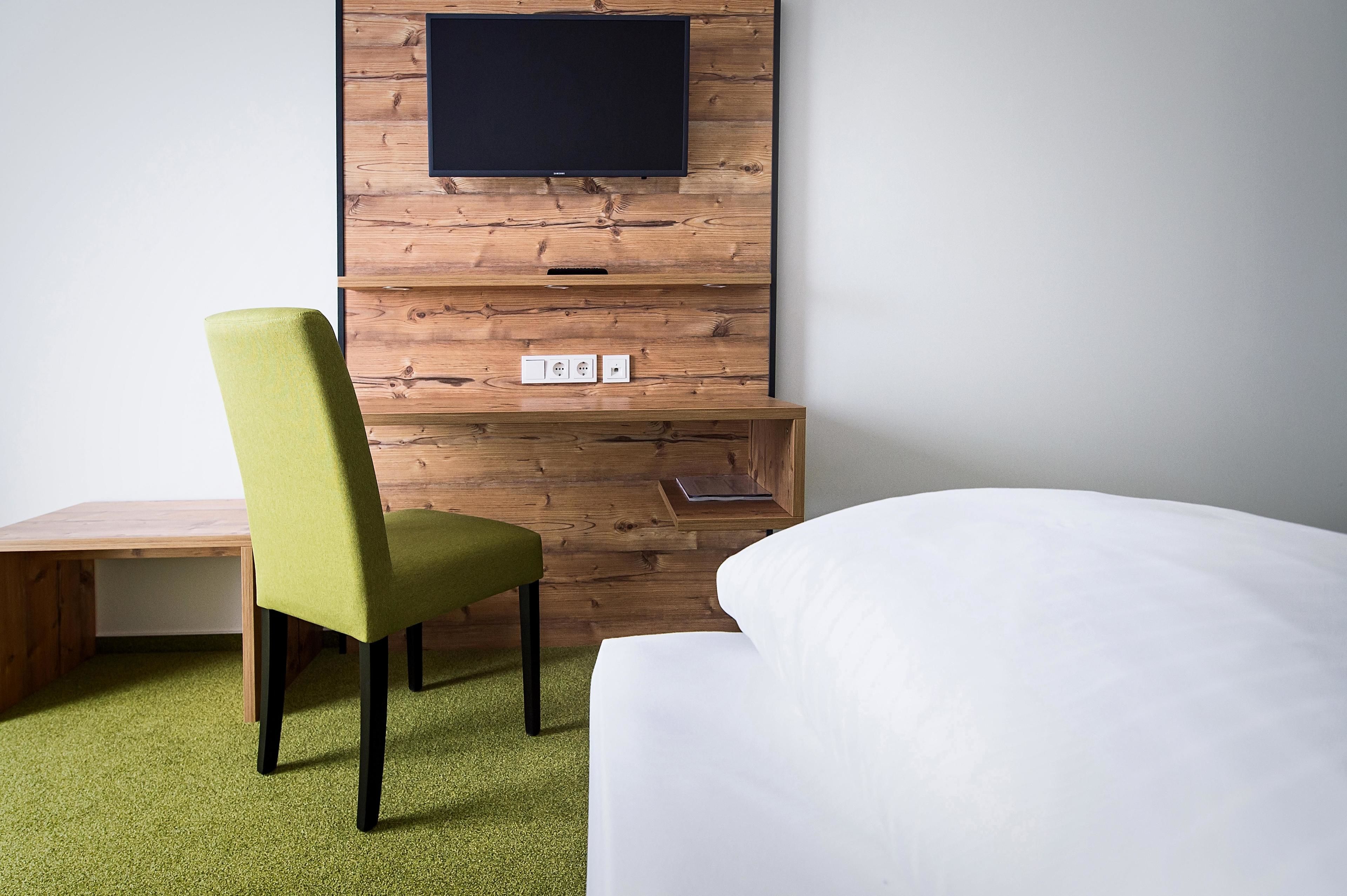 A hotel room with a wooden wall, a flat-screen TV mounted above a wooden shelf, a green upholstered chair, a wooden desk, and a white bed.