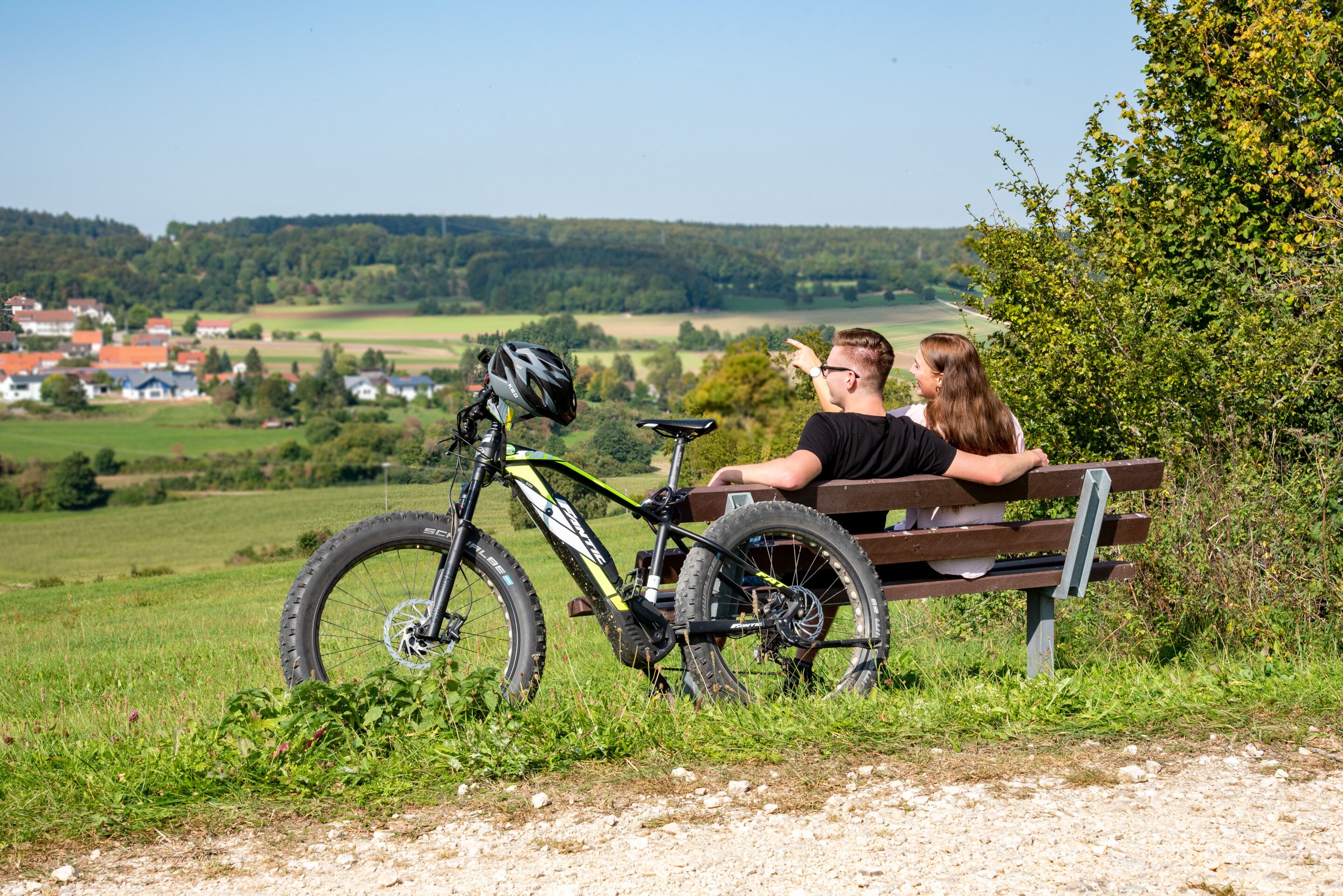 A couple sitting on a bench with a mountain bike parked beside them, overlooking a rural landscape with houses and fields.