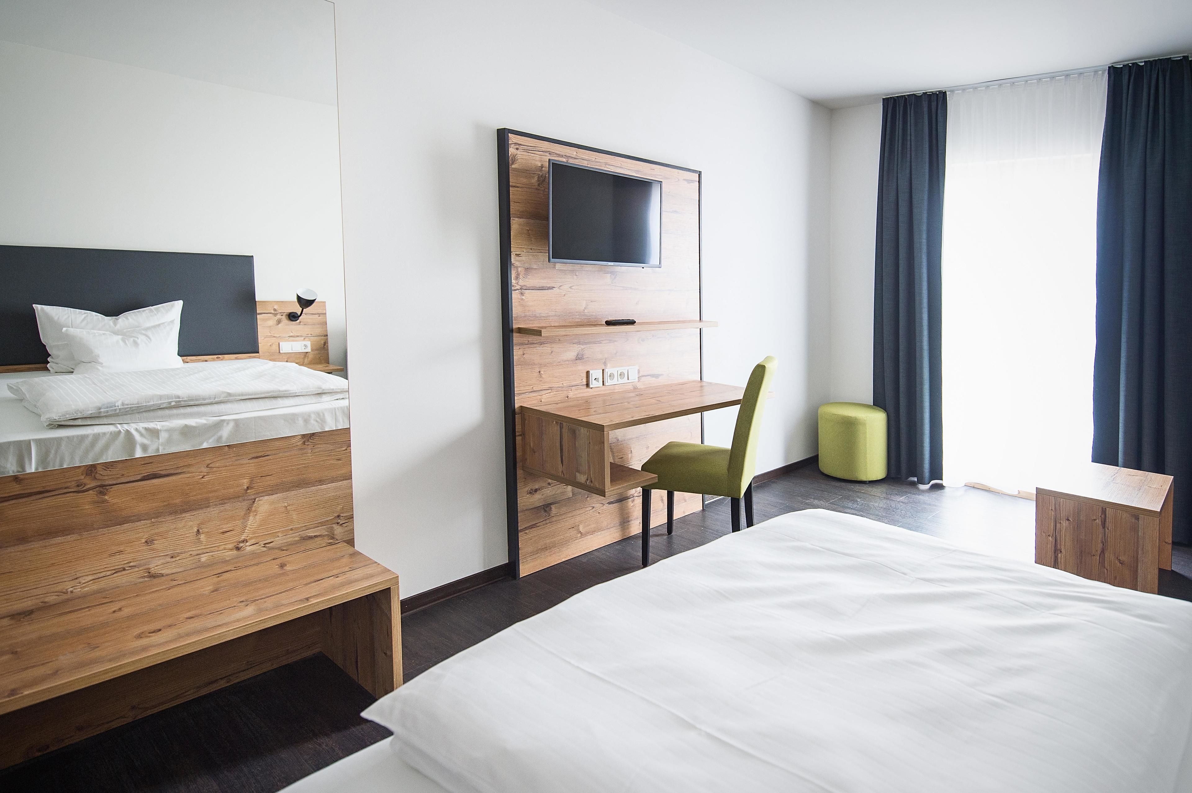 Habitación de hotel con cama, escritorio con silla, televisor montado en la pared y ventana con cortinas.