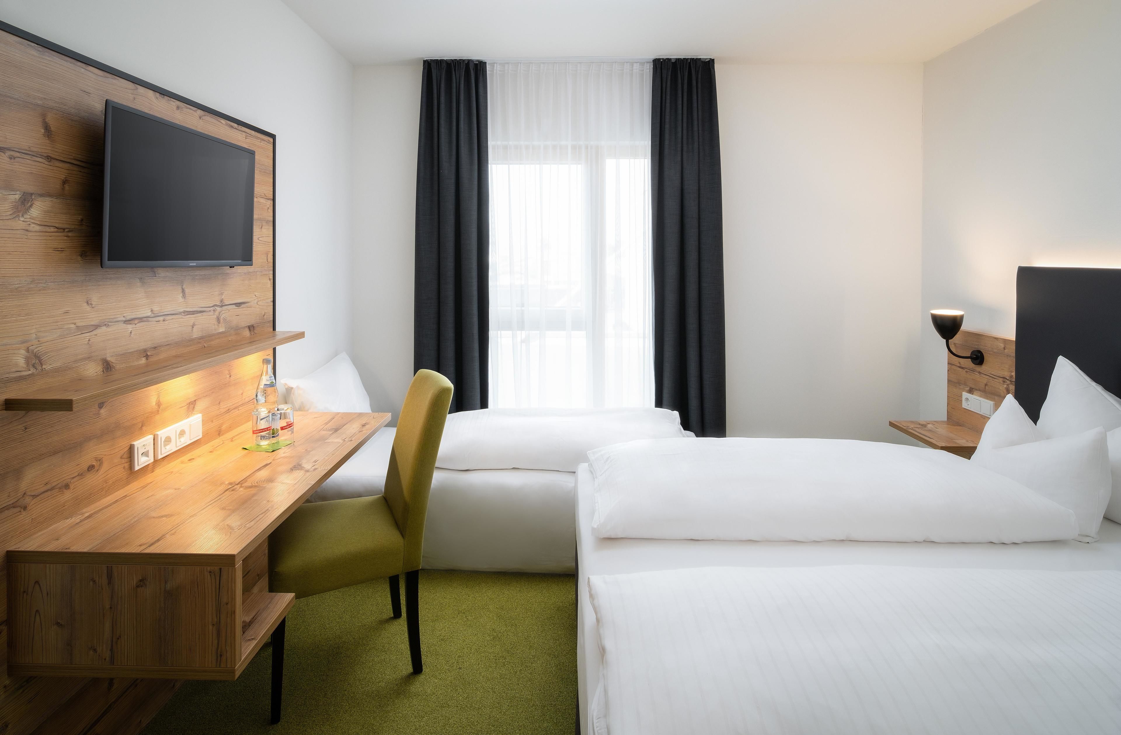 Habitación de hotel con un escritorio de madera, televisor montado en la pared, cama, silla y ventana con cortinas.