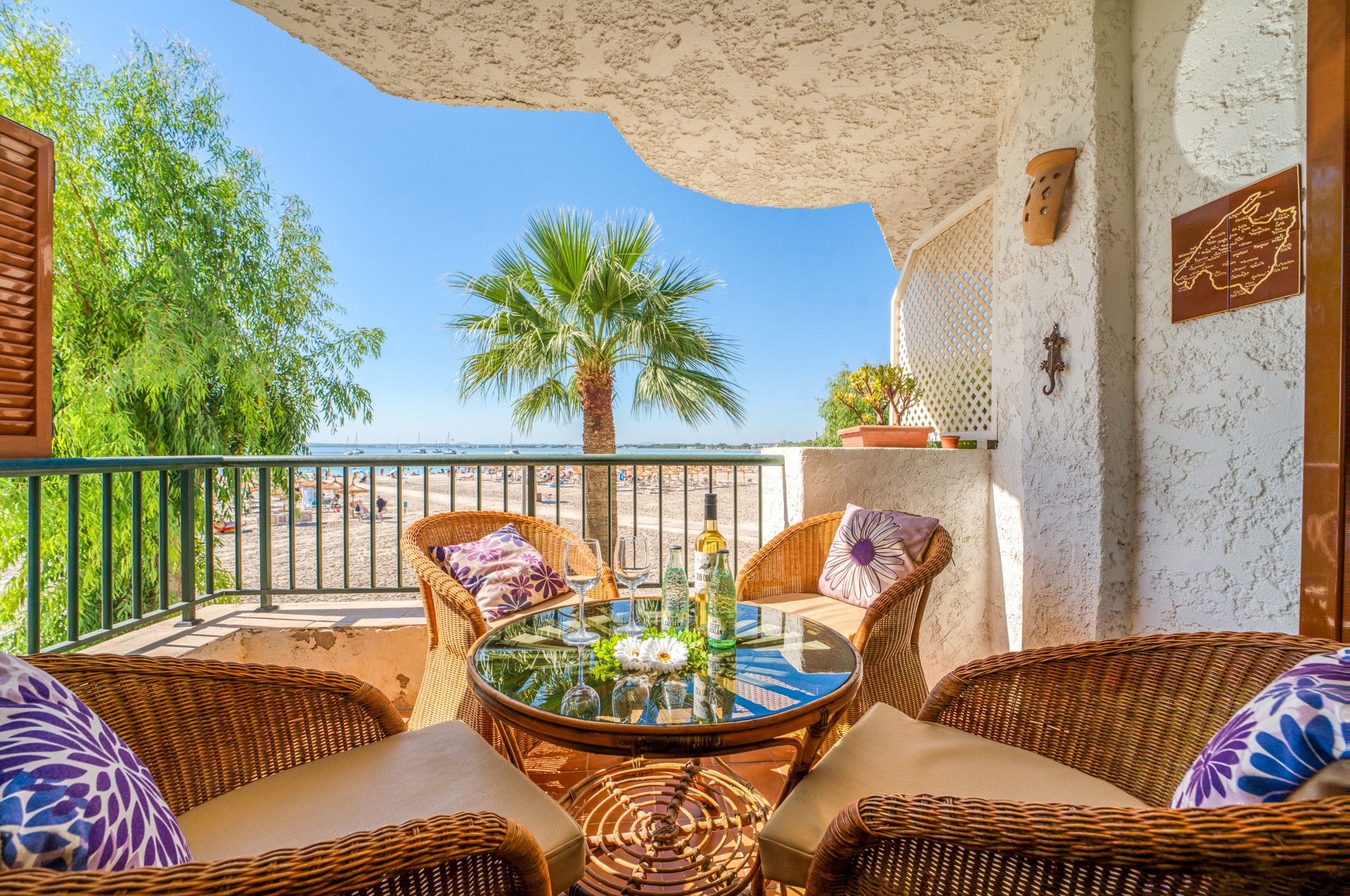 A balcony with wicker furniture, a glass table, and a view of the beach and palm trees.