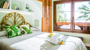 A bed with a white quilt, pillows with leaf patterns, a rolled towel with flowers, and a window with a view of greenery. - Apartment Carabela 44 on Alcudia Beach (Alcúdia)