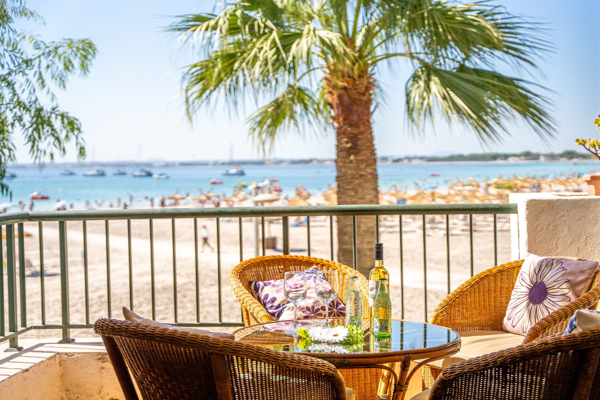 Une table en osier surmontée d’une plaque de verre, deux verres à vin et des bouteilles, le tout sur un balcon donnant sur une plage bordée de palmiers et de bateaux.
