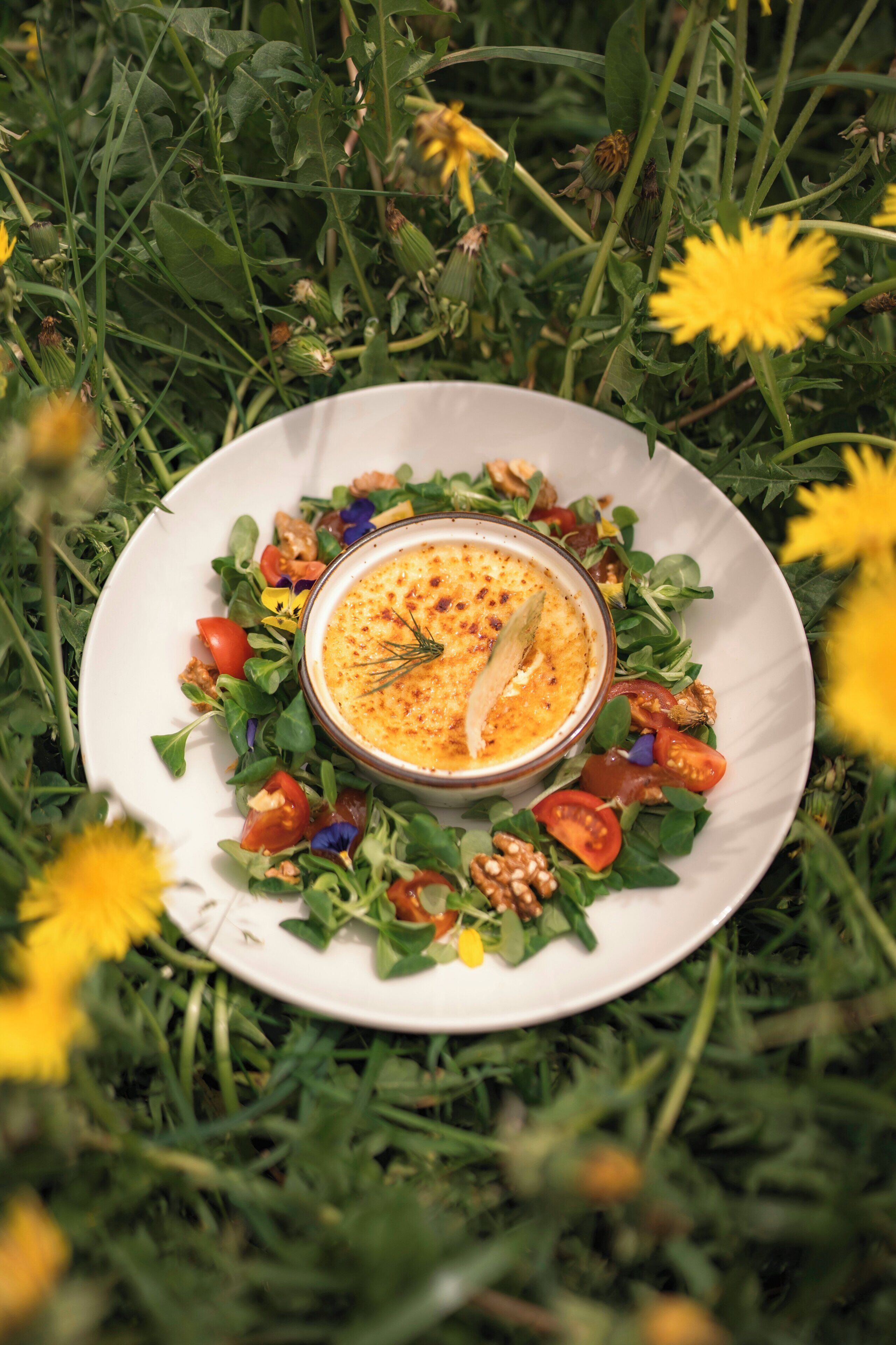 Un plato blanco con una salsa cremosa rodeado de ensalada, sobre pasto con dientes de león.