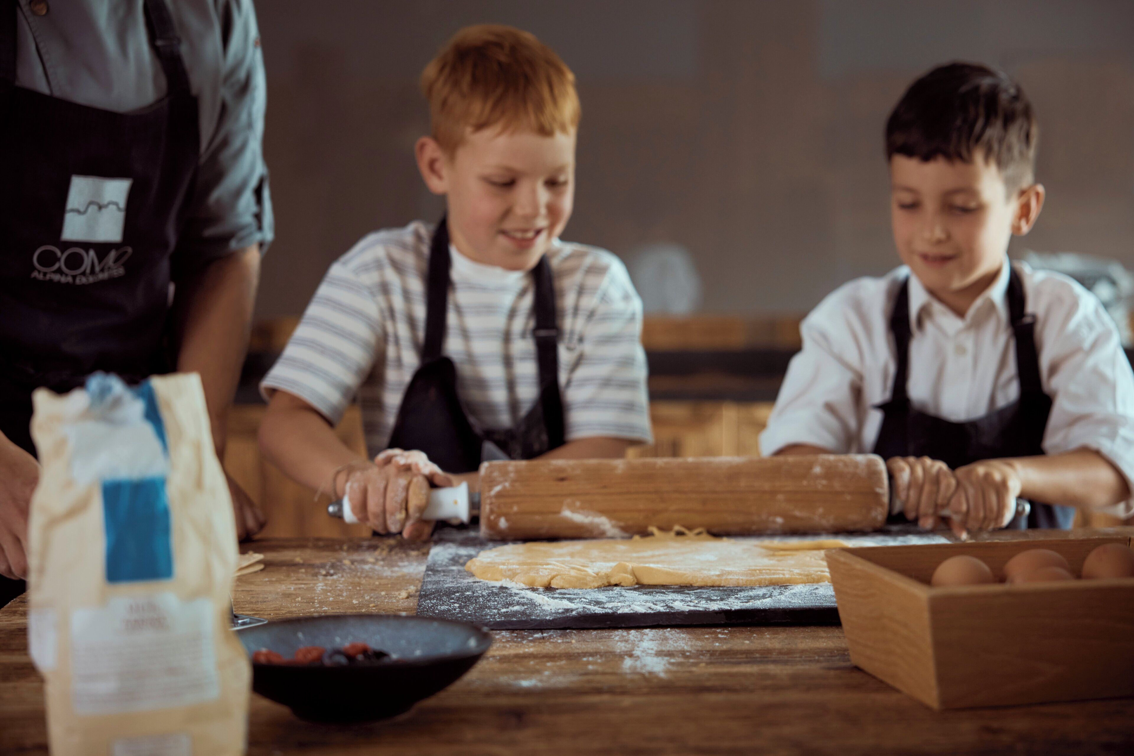 Escena de cocina con dos niños y un adulto, un rodillo, un bol de fruta y un saco de harina sobre la encimera.
