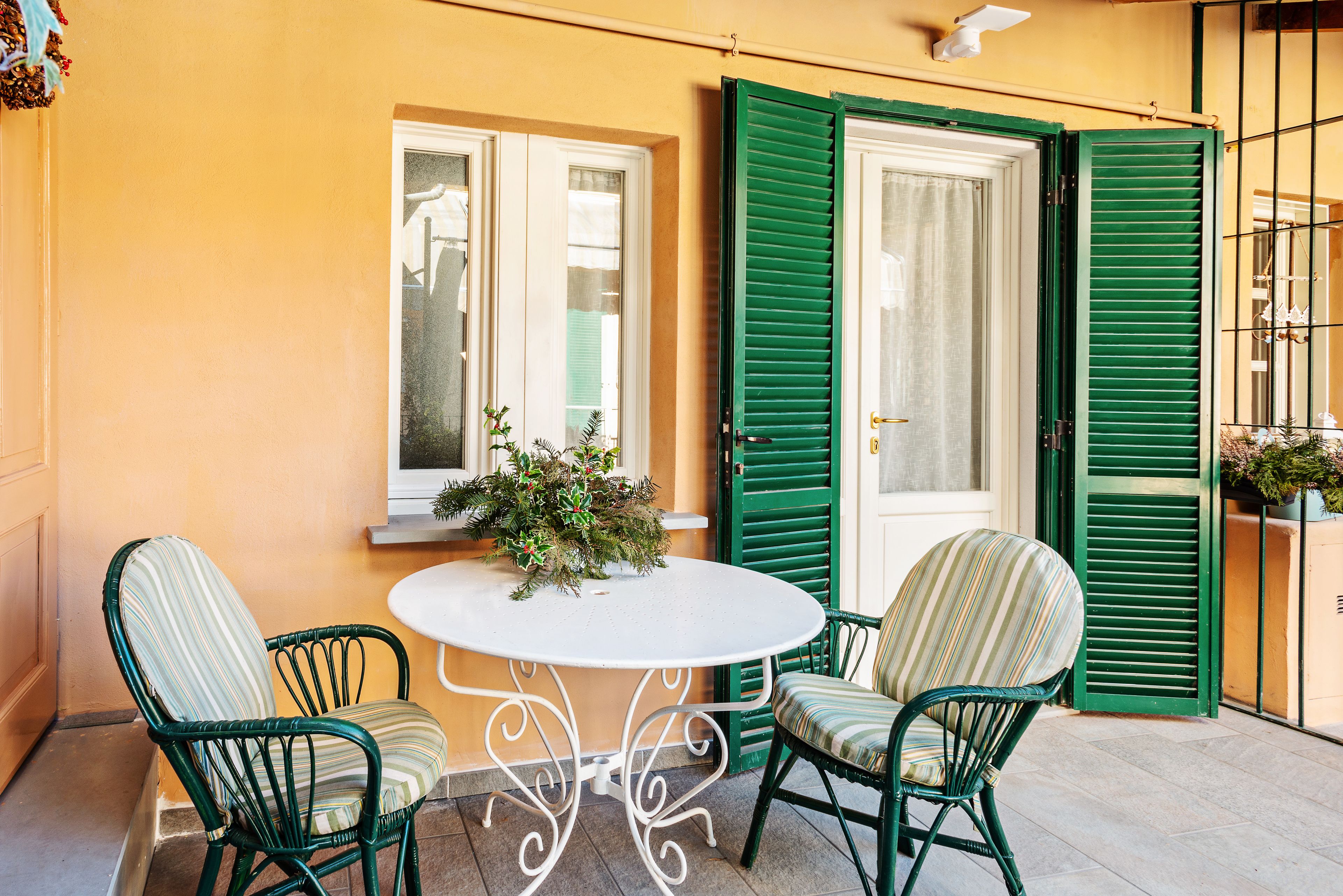 A small outdoor seating area with a round table and two chairs, a potted plant, and a window with green shutters.