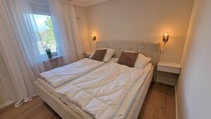 Een tweepersoonsbed met witte beddengoed, twee nachtkastjes met lampen, een raam met dunne gordijnen en uitzicht op groen.