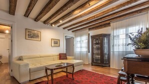 A living room with a white sofa, a red patterned rug, wooden beams on the ceiling, a cabinet with glass doors, and a potted plant. - Venezia - Calle Furlani 3277 (Venice)