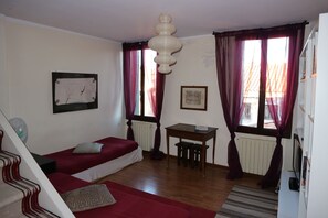 Una habitación pequeña y funcional con cama, escritorio y silla. Cuenta con ventana con cortinas, radiador y ventilador fijado a la pared.