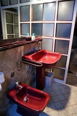 Un baño con lavamanos y bidé rojo, una ventana con paneles de vidrio esmerilado y suelo de baldosas.