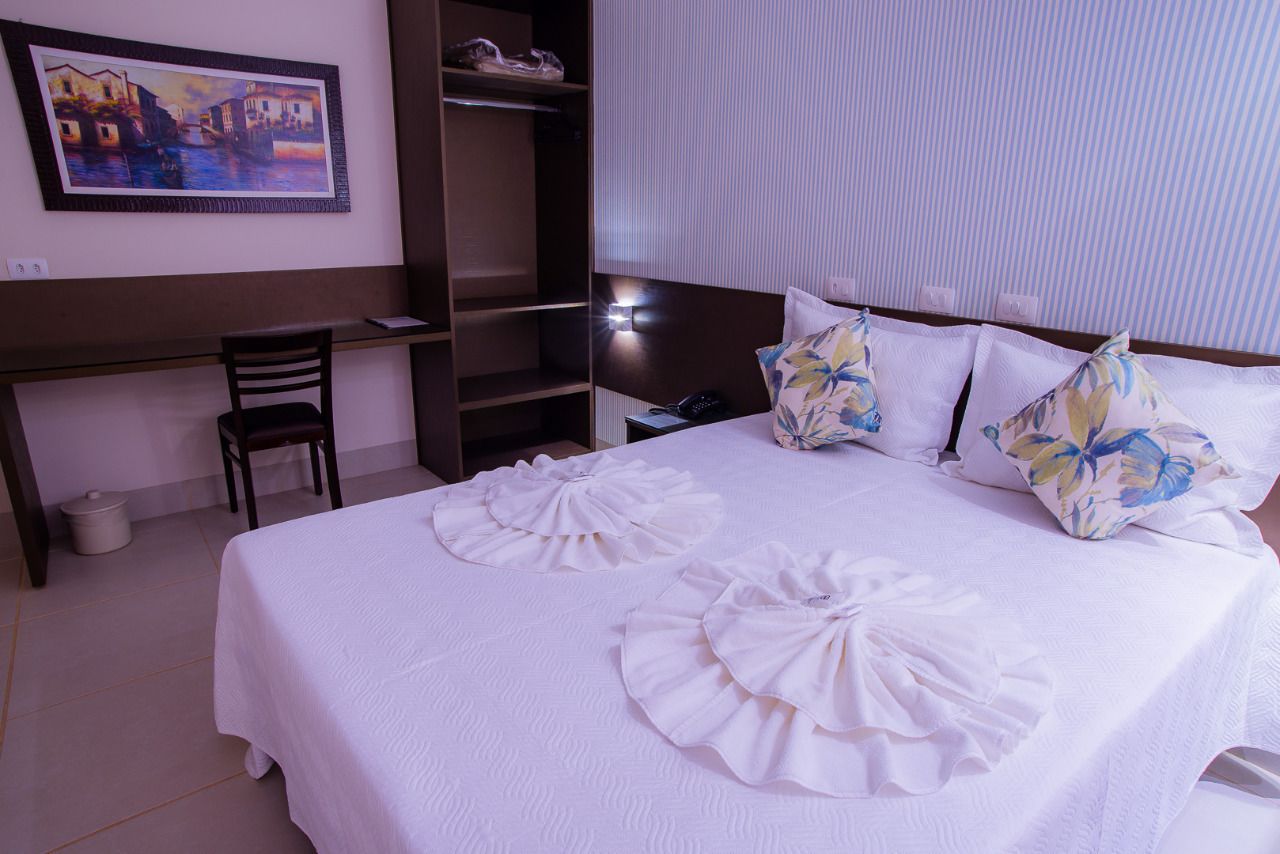 A hotel room with a large bed, a desk, and a chair. There is a painting on the wall and a window with blinds.