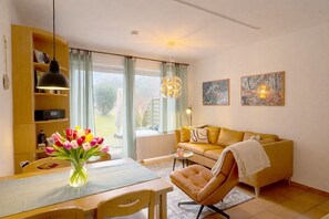 Un salon avec un canapé, une table à manger ornée d’un vase de fleurs, une bibliothèque et une fenêtre avec des rideaux.
