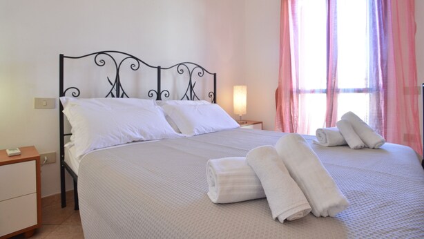 Una cama bien hecha con ropa blanca y toallas enrolladas, una mesita de noche con lámpara, y una ventana con cortinas rosadas.