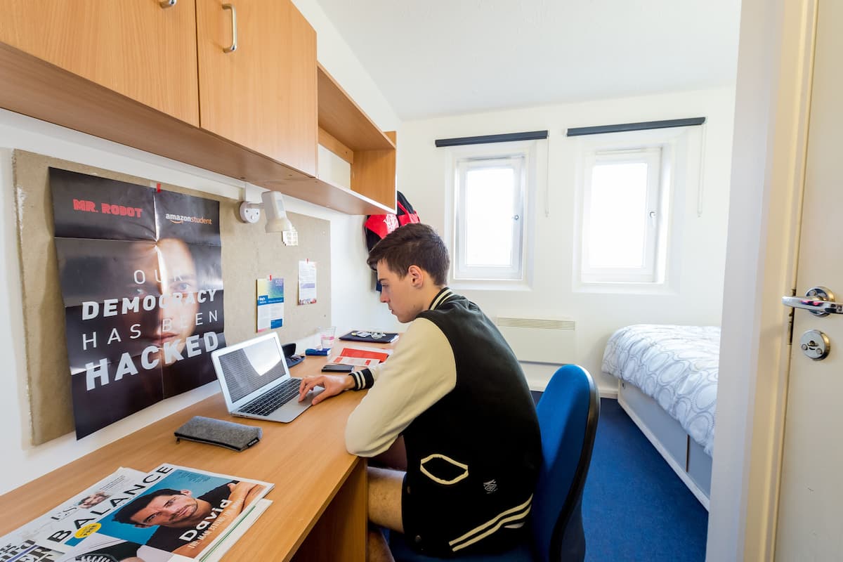 A room with a wooden desk, a laptop, a bed with a white and blue patterned duvet, and a poster on the wall.